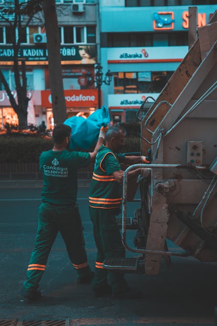 Two waste collection workers are actively engaged in rubbish removal on an urban street, standing next to a large industrial waste truck. The worker on the left, wearing a dark green uniform with refl