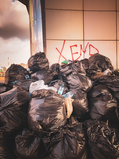 Two waste collection workers are actively engaged in rubbish removal on an urban street, standing next to a large industrial waste truck. The worker on the left, wearing a dark green uniform with refl