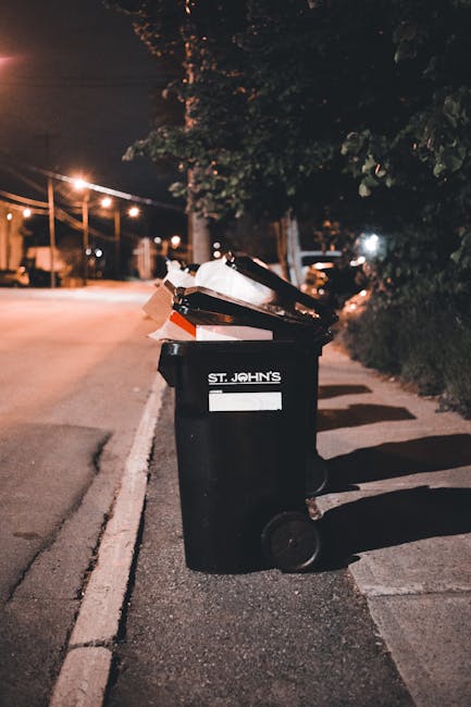 A black outdoor rubbish bin positioned along a paved sidewalk on a quiet street at night, illuminated by nearby streetlights. The bin is partially filled with various waste items, including paper and 