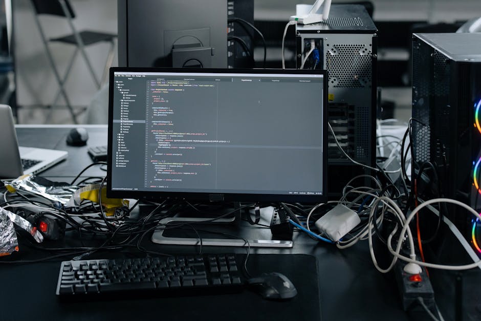 A close-up view of a person's hand operating a black computer keyboard on a desk, with a large computer monitor displaying lines of code or programming text on the left side of the image. The desk sur
